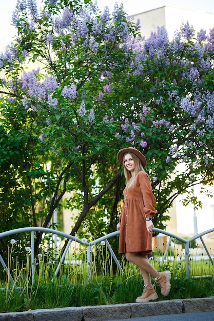 Retrato de niña linda con sombrero grande marrón y ropa elegante en el contexto de un árbol lila en un jardín botánico. Cabello largo castaño claro sobre los hombros.