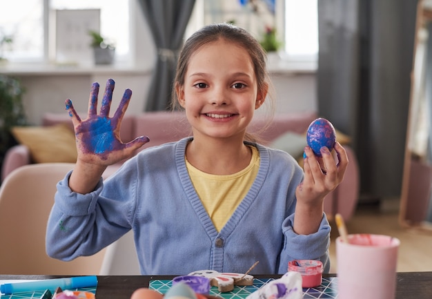 Retrato de niña linda sonriendo a la cámara mientras está sentado en la mesa con huevo de Pascua y mostrando su mano pintada