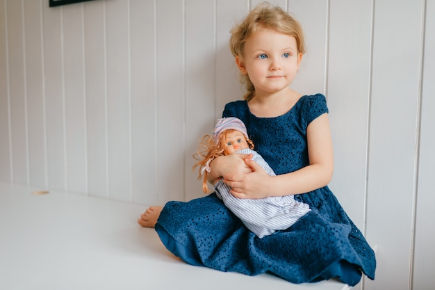 Retrato de niña linda sostiene a su encantadora barbie, se sienta en la habitación del bebé brillante, mira a un lado