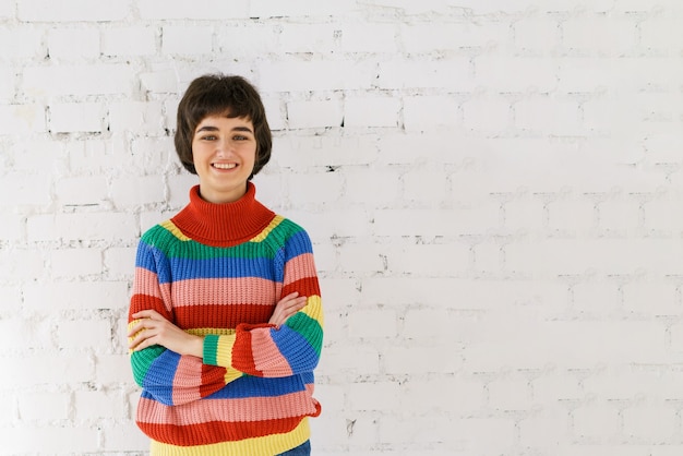 Retrato de niña linda en suéter de arco iris sobre fondo de pared de ladrillo blanco. Jóvenes mujeres caucásicas mirando a la cámara y sonriendo. Concepto LGBT.