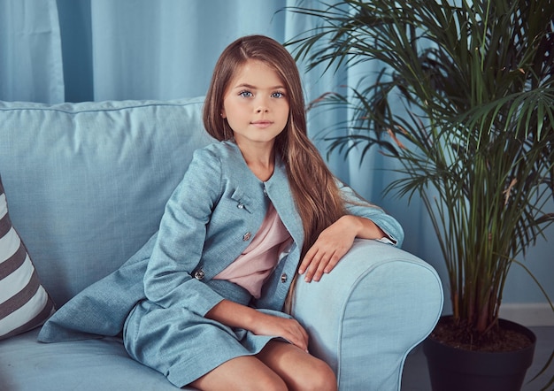 Retrato de una niña linda con un vestido elegante, sentada en un sofá en casa, mirando una cámara.