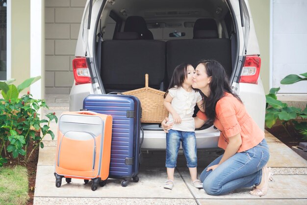 Retrato de una niña lista para viajar de vacaciones mientras besa a su madre cerca del coche, filmado en el garaje de la casa