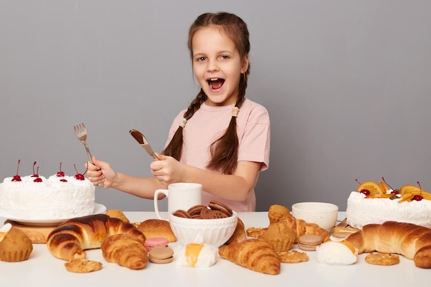 Retrato de una niña loca hambrienta y emocionada con coletas posando cerca de la mesa con deliciosos dulces sosteniendo un cuchillo y un tenedor listos para comer pastel de cumpleaños aislado sobre fondo gris