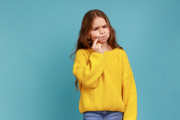 Retrato de una niña malsana que toca la mejilla sintiendo dolor de muelas severo problemas de salud oral usando un suéter amarillo de estilo casual Foto de estudio interior aislada en fondo azul