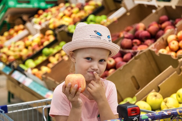 Retrato de niña con manzana sentada en un carrito de compras en una tienda de frutas pensando profundamente y mirando a la cámara