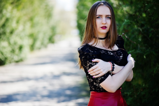 Retrato de niña con maquillaje brillante con labios rojos, collar de gargantilla negra en el cuello y falda de cuero rojo.