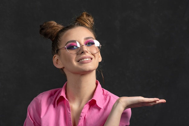 Retrato de niña con maquillaje de noche en gafas con peinado de moño en camisa rosa fondo negro Lugar para el texto