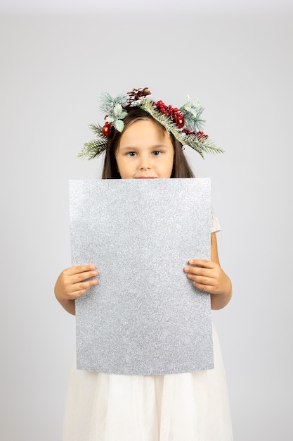 Retrato de niña maravillosa en vestido blanco y corona de navidad sosteniendo cartel plateado brillante aislado ...