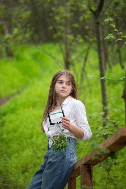 Retrato de una niña de moda, de pie apoyado contra un puente de madera de la aldea, con el telón de fondo de los árboles verdes del bosque.