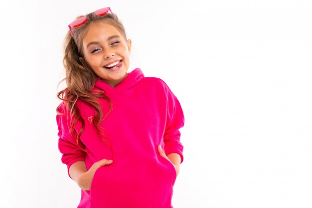 Foto retrato de niña de moda en sudadera rosa, gafas de sol rosadas es feliz y tiene las manos en los bolsillos, aislado en blanco