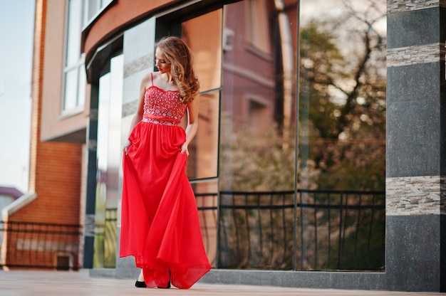 Retrato de niña de moda en el vestido de noche rojo que plantea la ventana de espejo de fondo del edificio moderno