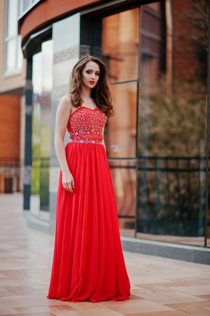 Retrato de niña de moda en el vestido de noche rojo que plantea la ventana de espejo de fondo del edificio moderno