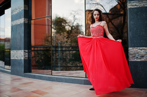 Retrato de niña de moda en el vestido de noche rojo que plantea la ventana de espejo de fondo del edificio moderno