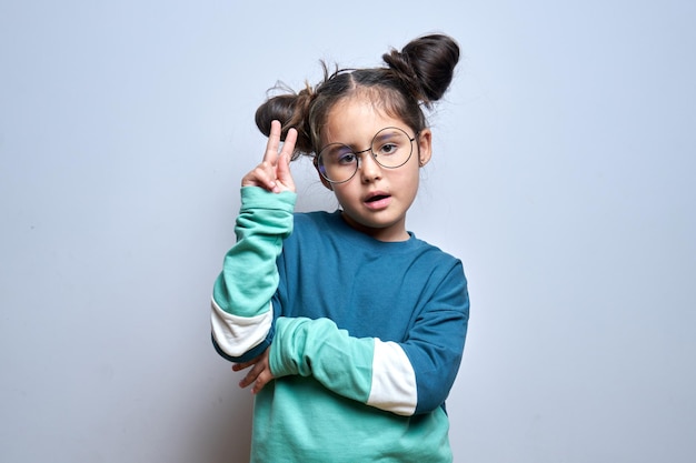 Retrato de niña morena positiva mostrando gesto de paz en vasos redondos aislado sobre fondo blanco.