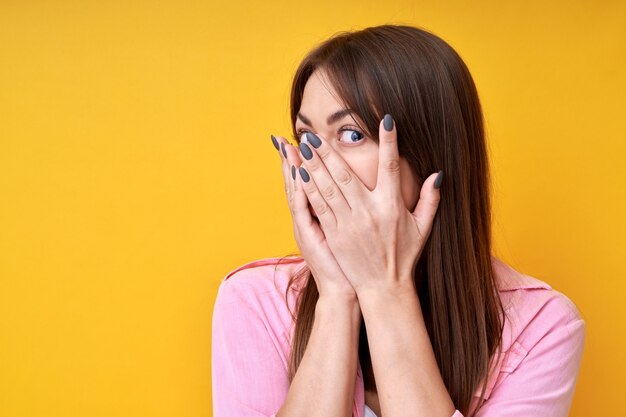 Foto retrato de niña morena que cubre sus ojos aislados sobre fondo amarillo, mirando a través de los dedos, mirando furtivamente a la cámara, con miedo de mirar