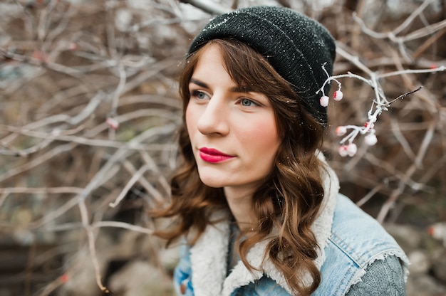 Retrato de niña morena con sombrero y chaqueta de jeans en arbustos congelados.