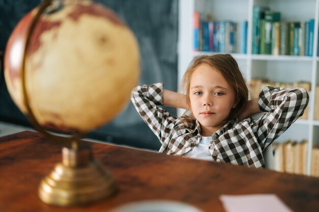 Retrato de niña niño curioso sentado en una silla con las manos detrás de la cabeza delante de la mesa con un