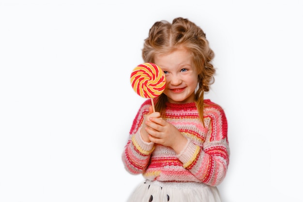 Retrato de una niña un niño rubio con una paleta en un palo sonriendo en ropa de colores sobre un fondo blanco.