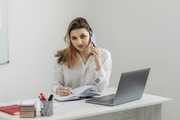 Retrato de una niña en la oficina La niña está hablando por teléfono y trabajando en una computadora portátil Escribe en un cuaderno La niña está vestida con una camisa blanca El concepto de trabajo Enseñanza