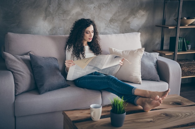 Retrato de niña pacífica concentrada sentada en el sofá leyendo noticias de finanzas en todo el mundo en la moderna sala de estar de estilo interior de loft industrial en el interior