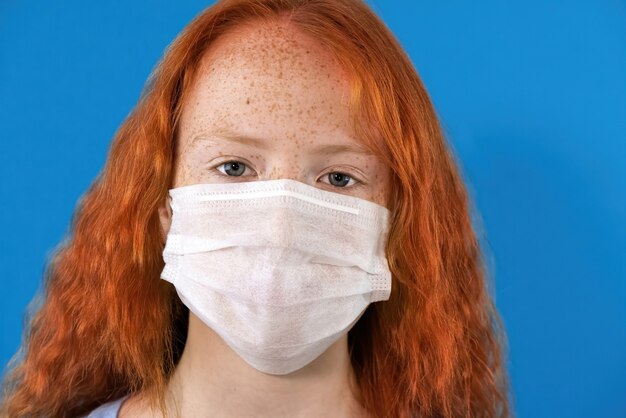 Retrato de una niña pelirroja con una máscara médica blanca con ojos azules y pecas en azul