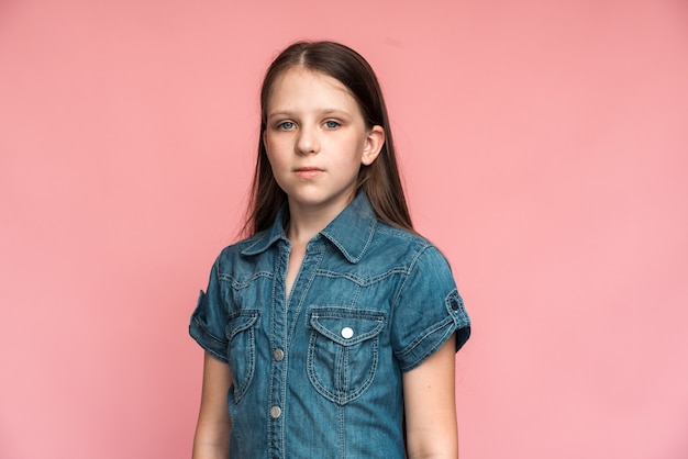 Retrato de niña de pelo largo preescolar feliz en camiseta de jeans tranquilamente mirando a cámara y sonriendo, orgullosa de su propio éxito. Tiro de estudio interior aislado sobre fondo rosa