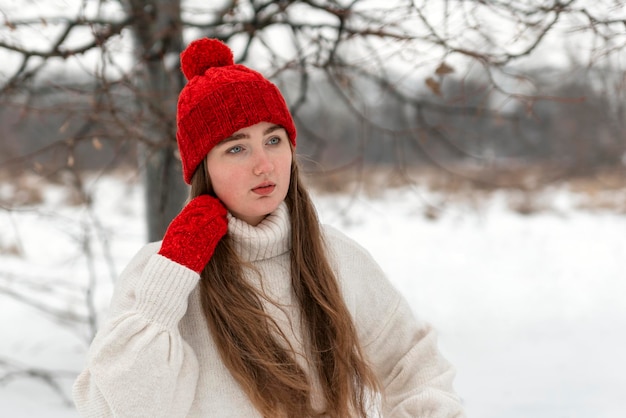 Retrato de niña de pelo largo con sombrero rojo de punto de pie en un parque cubierto de nieve Mujer en ropa de abrigo en invierno al aire libre