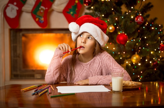 Retrato de niña pensativa en gorro de Papá Noel pensando en carta con deseos de regalo
