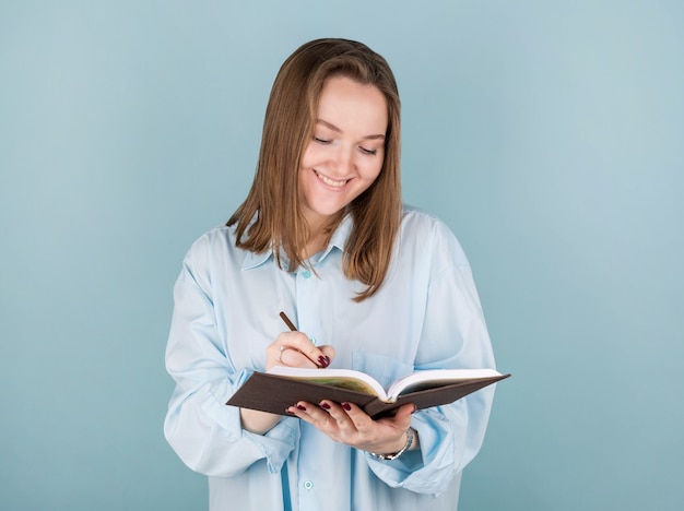 Retrato de niña pensativa pensativa con cuaderno y lápiz en la boca. Aislado en azul con espacio de copia.