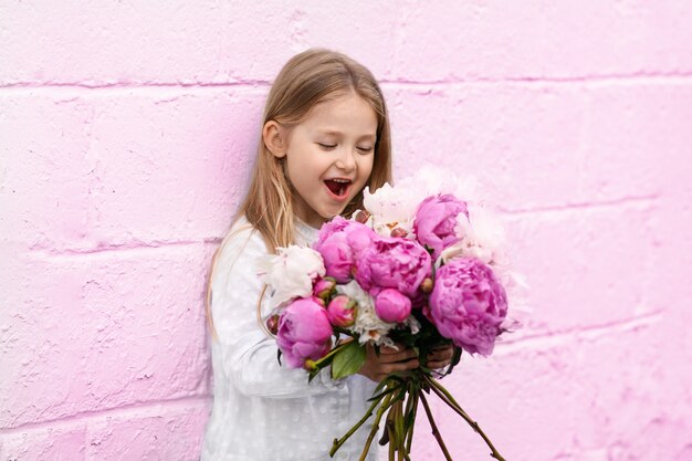 Retrato de una niña con las peonías cerca de la pared rosa