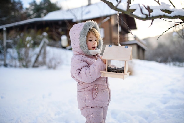 Retrato de niña pequeña al aire libre en el jardín de invierno, de pie junto al comedero para pájaros de madera.