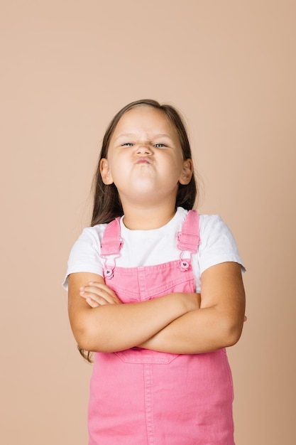 Retrato de una niña pequeña con los brazos cruzados con cara de mueca insatisfecha mirando a la cámara con mono rosa brillante y camiseta blanca sobre fondo beige