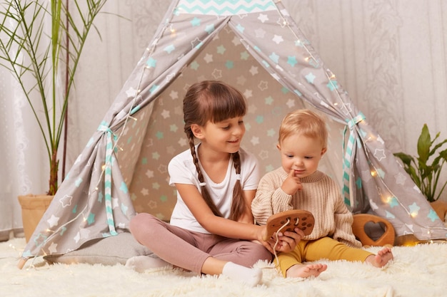 Retrato de una niña pequeña con coletas con una camiseta blanca jugando con juguetes de madera en el wigwam junto con su hermana pequeña cuidando a un niño pequeño que tiene un ventilador en una tienda tipi