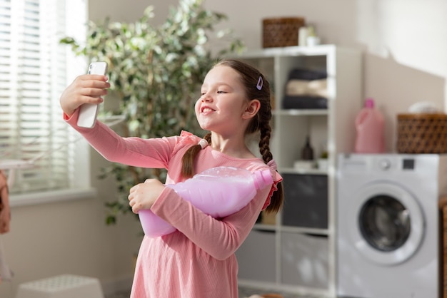 Retrato de una niña pequeña con coletas con un vestido rosa. El niño sostiene un teléfono inteligente hablando por teléfono grabando un video en las redes sociales. En la otra mano, sostiene un líquido para lavar la ropa.