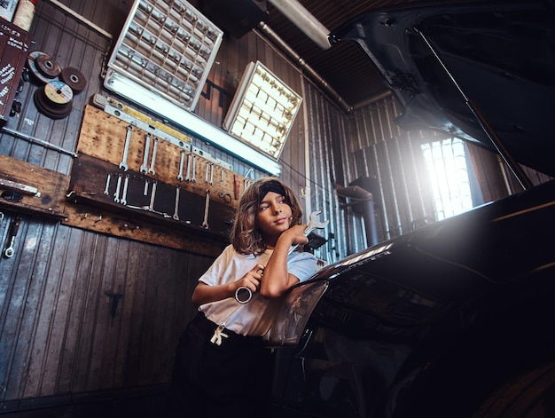 Retrato de una niña pequeña y hermosa con una gran herramienta en las manos. Ella está parada cerca del auto en el taller de autos.