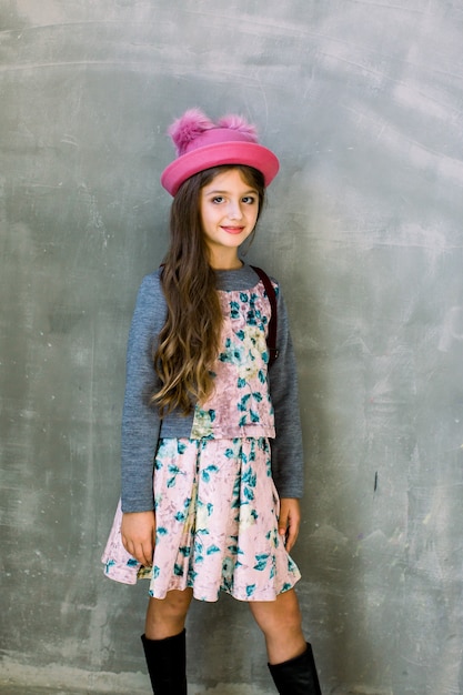 Retrato de niña pequeña de moda con gorra rosa y vestido con flores y una mochila marrón en una pared gris