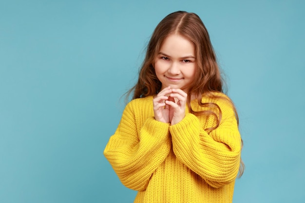 Retrato de una niña pequeña pensando en una idea ingeniosa y sonriendo a la cámara, teniendo planes complicados en mente, usando un suéter amarillo de estilo casual. Disparo de estudio interior aislado sobre fondo azul.