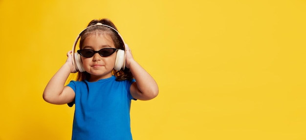 Retrato de niña pequeña posando en auriculares y gafas de sol de moda aislado sobre fondo amarillo Concepto de emociones de moda de verano infantil Flyer