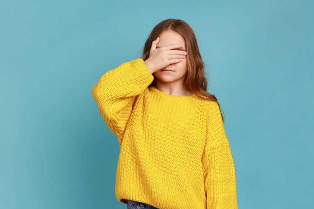 Retrato de una niña pequeña que cubre los ojos con la mano para no mirar algo prohibido ocultar los ojos usando un suéter amarillo de estilo casual Foto de estudio interior aislada en fondo azul