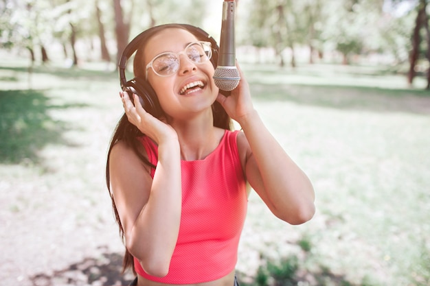 Retrato de niña de pie afuera y cantando al micrófono. Ella está escuchando música al mismo tiempo mientras canta. Joven mujer está disfrutando el momento.