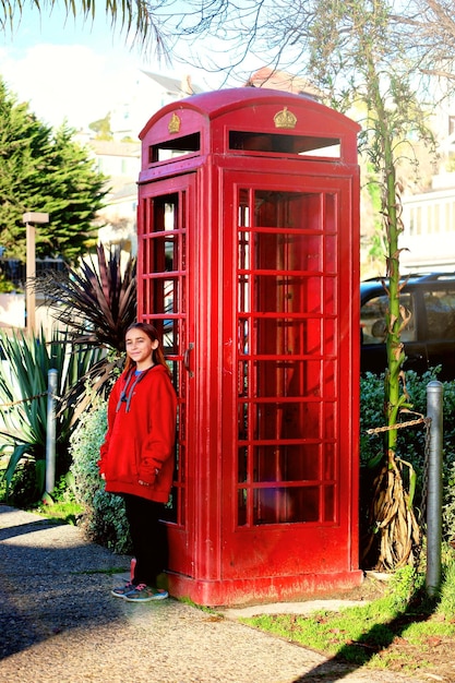 Foto retrato de una niña de pie contra una cabina telefónica