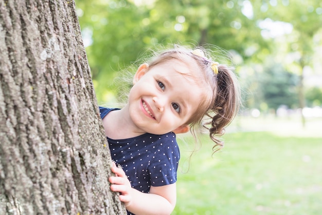 Retrato de una niña de pie detrás del árbol mirando a escondidas en el jardín