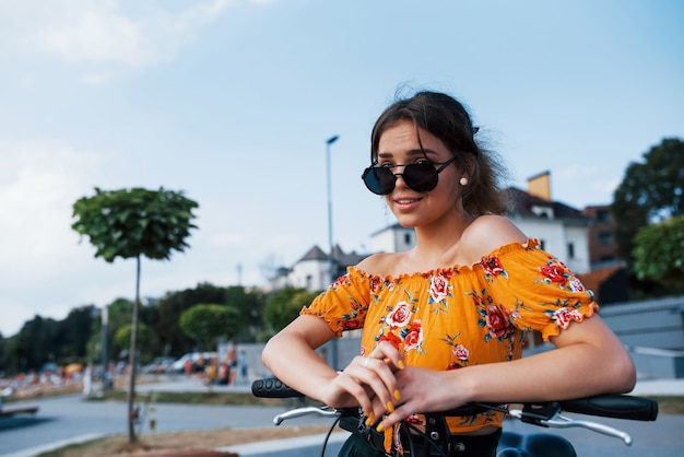 Retrato de niña positiva en gafas de sol que se encuentra con su bicicleta.