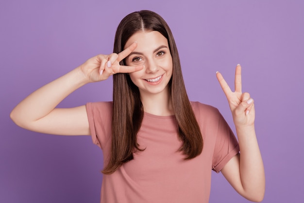 Retrato de niña positiva gesticulando dos signos v cerca de la cara aislada sobre fondo violeta