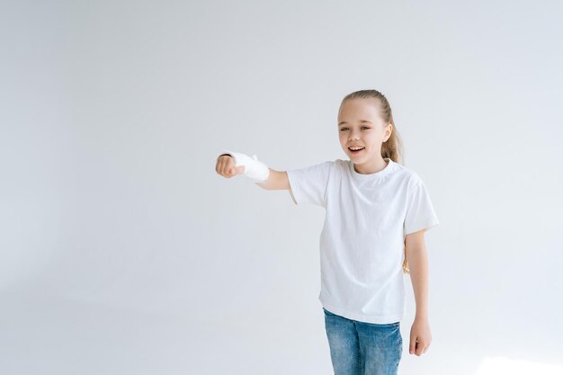 Retrato de una niña positiva con la mano rota envuelta en una venda de yeso blanco jugando gesticulando con la muñeca lesionada en un fondo claro y aislado