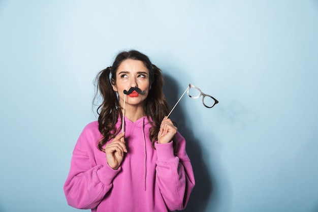 Retrato de niña princesa feliz con bigote de papel y gafas de sol falsas en palo sobre azul en estudio