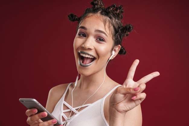 Retrato de niña punk alegre con peinado extraño y lápiz labial oscuro que muestra los dedos de la paz mientras usa un teléfono inteligente con auriculares aislados sobre una pared roja