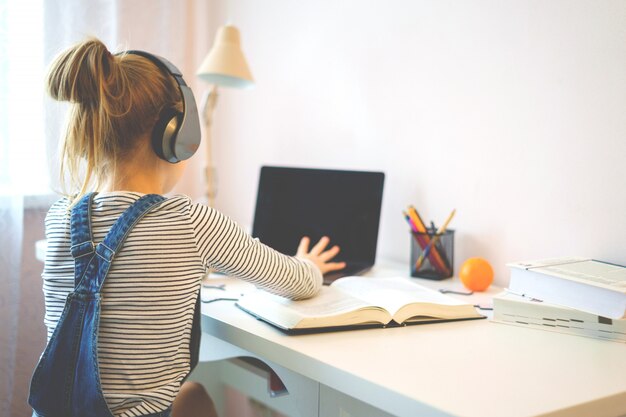 Retrato de una niña que aprende en línea con auriculares y computadora portátil tomando notas en un cuaderno sentado en su escritorio en casa haciendo la tarea