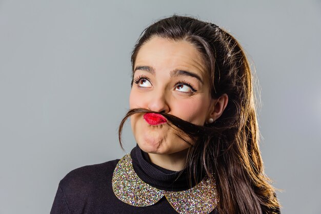 Retrato de niña real con labios rojos usando su cabello como un gracioso bigote sobre un fondo gris