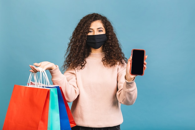 Retrato de niña rizada hermosa emocionada en una máscara protectora médica en su rostro, sosteniendo bolsas de compras aisladas sobre un fondo rosa